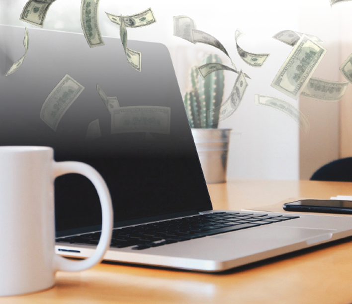 Money falling on top of computer sitting on desk with coffee cup near by